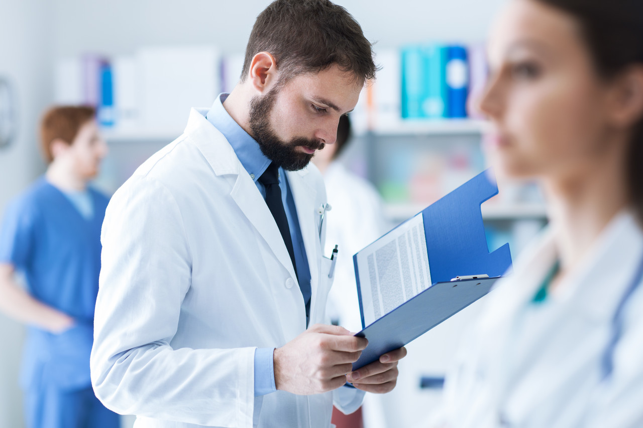 Confident doctor checking medical records on a clipboard in his office, medical staff on the background, healthcare concept